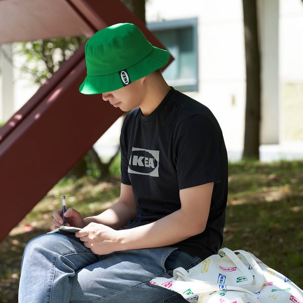 A young person sits on grass, wearing a green bucket hat and black t-shirt, writing in a notebook. Beside them, a colorful tote bag.