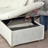 Person lifting white sofa ottoman lid, revealing storage space lined with foam for comfort and organization, highlighting its practical design.