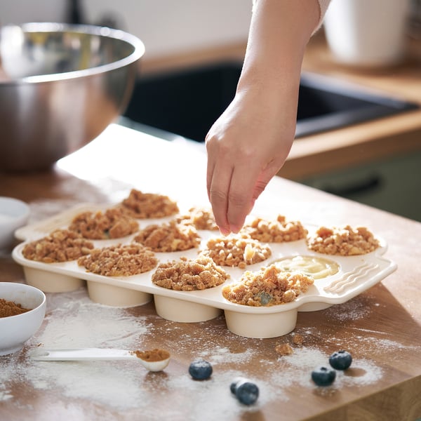 Hand placing crumble on muffin tray on wooden board with scattered blueberries.