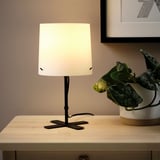 A small, modern lamp on a wooden table. The lampshade is lit, providing soft, glare-free light, enhancing room ambiance. A plant sits beside it.