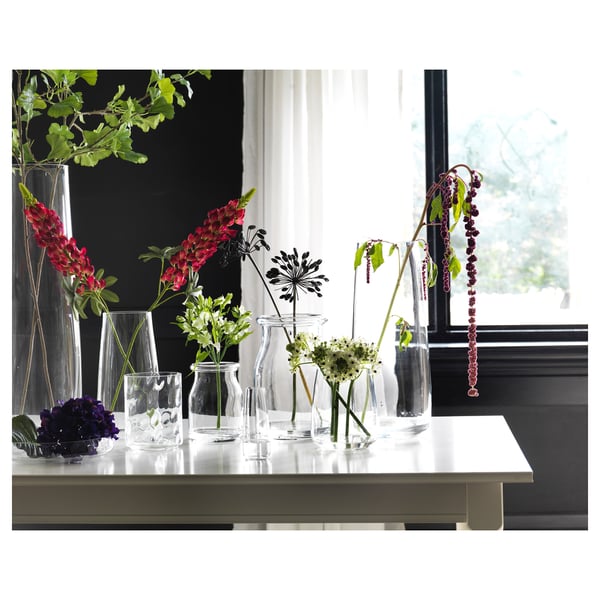 Several colorful flowers in clear glass vases on a white table near a window.