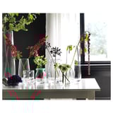 Several colorful flowers in clear glass vases on a white table near a window.