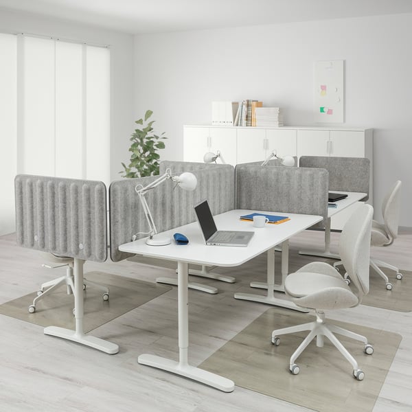 Modern office with adjustable BEKANT desk, laptop, supplies, gray chairs, privacy screens, large windows.