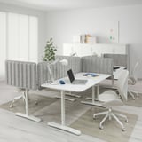 Modern office with adjustable BEKANT desk, laptop, supplies, gray chairs, privacy screens, large windows.