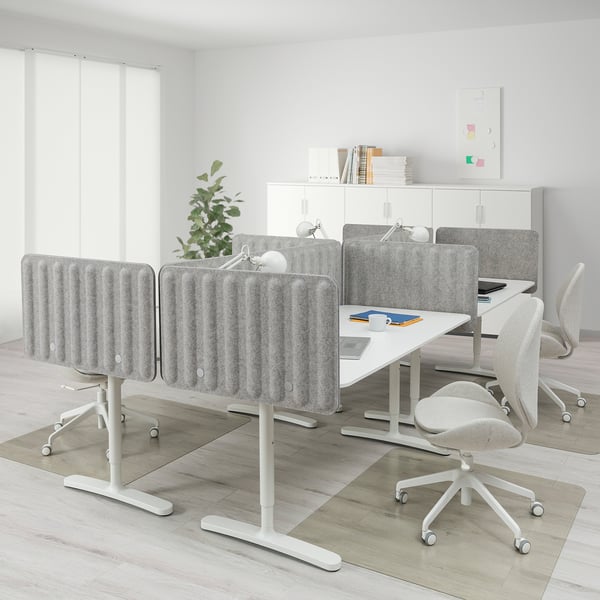 This image shows a modern office setup with a BEKANT white desk and light gray acoustic panels, larger than the surrounding light beige office chairs.