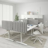 This image shows a modern office setup with a BEKANT white desk and light gray acoustic panels, larger than the surrounding light beige office chairs.