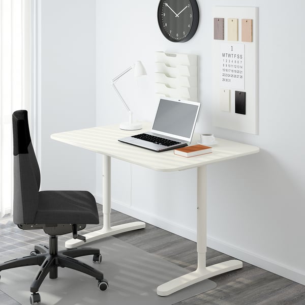 A minimalist home office setup with an adjustable BEKANT desk, laptop, lamp, chair, wall clock, and calendar.