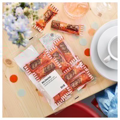 Opened chocolate bars BELÖNING on wooden table with wine glasses, plate, cup, flowers, confetti.