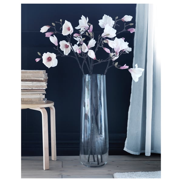 Clear glass BERÄKNA vase with white/pink magnolias by window, books, stool.