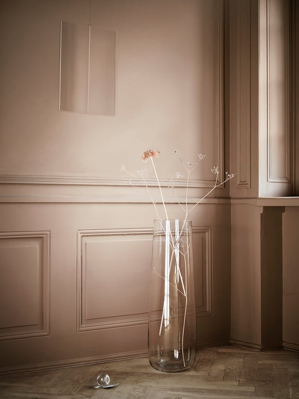 Clear glass vase with pale flowers on wooden floor, beige panelled wall, minimalist decor.