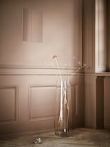 Clear glass vase with pale flowers on wooden floor, beige panelled wall, minimalist decor.
