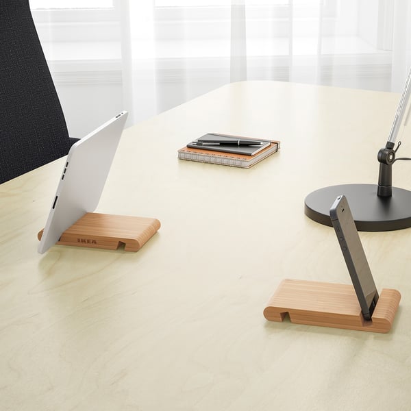A wooden stand holding a laptop and a tablet upright on a desk. The table is also holding a notepad, pens, and a monitor.