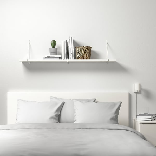 Minimalist bedroom: white BERGSHULT shelf holds books, plant, basket; white bed with pillows. Shelf is large compared to items.