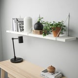 White wall shelf holding books, vases, and a plant with a black adjustable desk lamp beside a light wood desk.