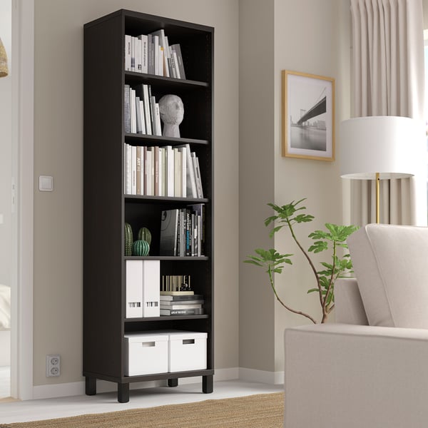 Black BESTÅ bookshelf with books, decor, by window, chair, lamp, plant.