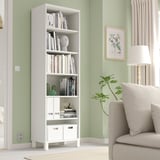 White BESTÅ bookcase against green wall, holding books and decor. Beige armchair, floor lamp, and plant nearby.