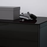 Black gaming controller resting on sleek, glossy glass-topped furniture, beside grey box.