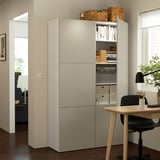 Modern home office with beige BESTÅ storage unit. Shelves adjustable, door open showing organized items. Desk with chair, lamp, and potted plant.