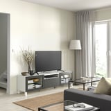 Modern living room with BESTÅ tv stand, black tv, glass doors, wooden legs, white walls, large window with curtains, rug, and black sofa.
