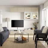 Modern living room with grey sofa, glass table, BESTÅ tv bench, beige cabinets, flat screen tv, and floral arrangement.