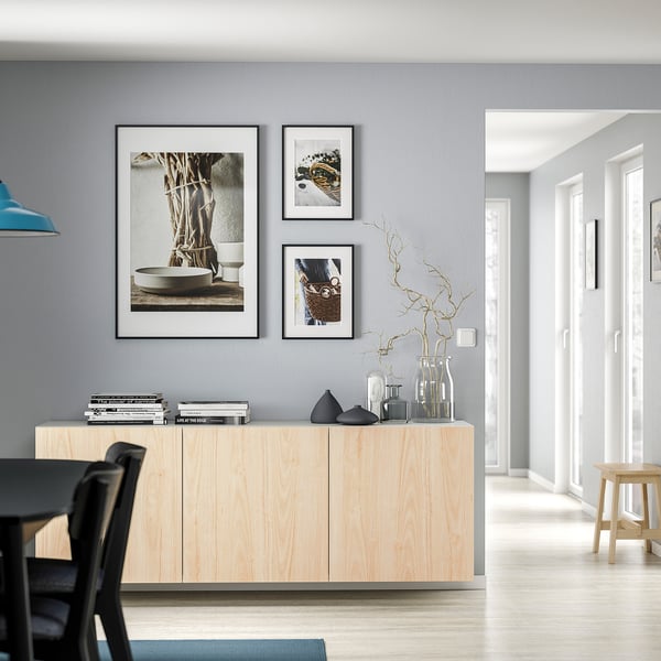 Minimalist living room with light wood BESTÅ cabinet, black accents, vases, books, framed pictures.