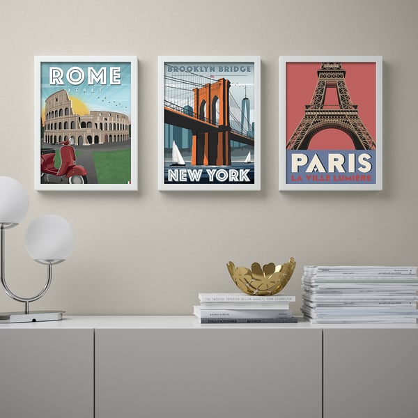 Three posters of rome, brooklyn bridge, and paris hang above a grey sideboard. The sideboard holds a white lamp, a gold bowl, and stacks of books.