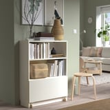 White BILLY bookcase with wheels holding books & vases. Smaller than couch, next to wooden stool.