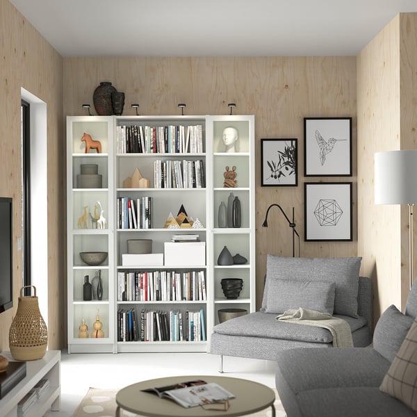 Modern living room with white BILLY bookcase, gray sofa, coffee table, and wooden walls. Decor includes books, vases, and framed art.