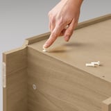A hand installs a white hinge on the side of a BILLY beige wooden bookcase.