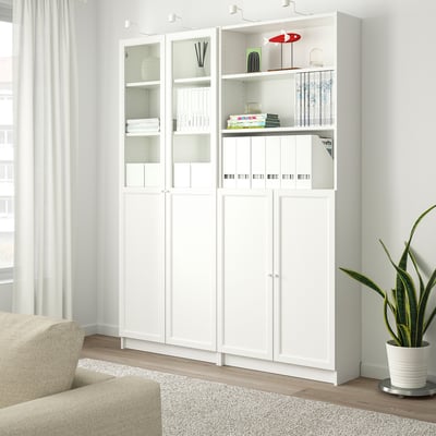White BILLY bookcase with glass doors, holding books and decor, next to a beige chair and a large potted plant.