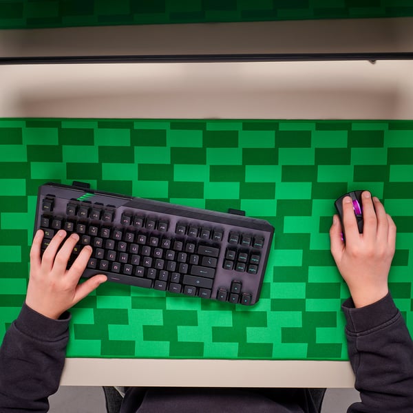 Person using keyboard and mouse on green checkered desk mat. Hands on keys and mouse, mouse pad helps smooth mouse control.