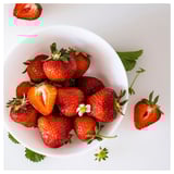 White bowl with red strawberries, some sliced, green leaves, and flowers.