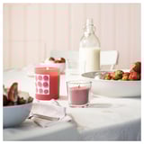 Table scene with red & purple candles, fresh strawberries, bowls, and a bottle. Light pastel background.