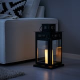 Black BORRBY lantern on floor next to white sofa, glowing candle inside, providing warm light.