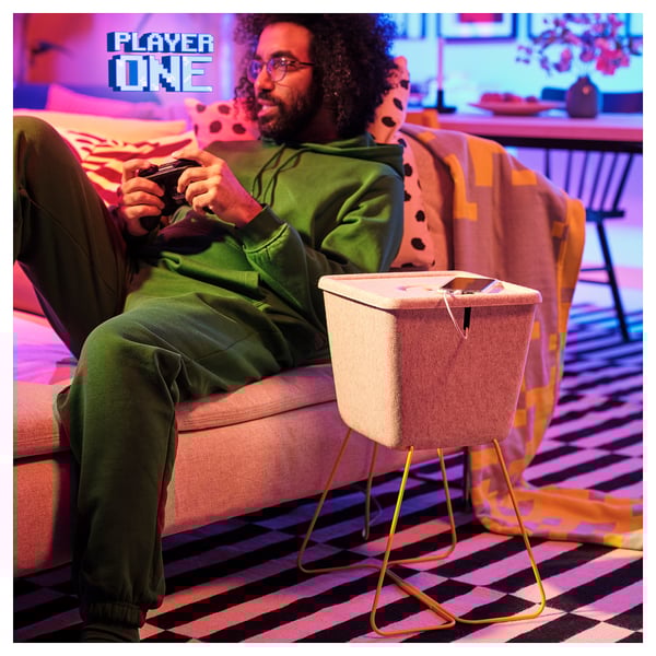 Man sits on pink couch, playing a game with remote. Beside him, a beige felt basket with gold legs holds a phone for charging.