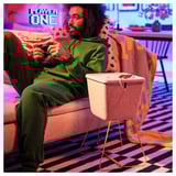 Man sits on pink couch, playing a game with remote. Beside him, a beige felt basket with gold legs holds a phone for charging.