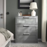 Modern grey BRIMNES chest of drawers with frosted glass top, white lamp, makeup items, mirror, and white bed in a small room.