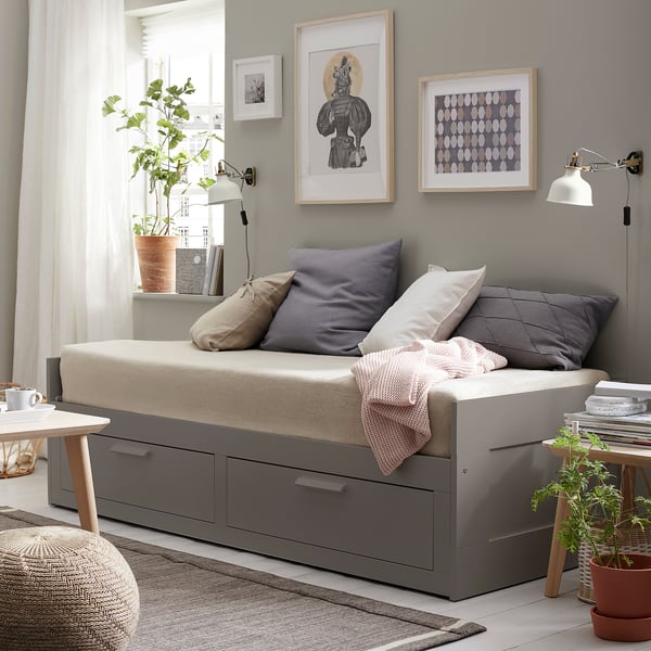 Dark grey day-bed with pillows, storage drawers, side tables with plants, and wall art in a light, minimalist room.