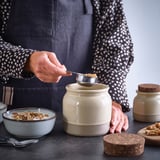 A woman scoops granola from a rustic stoneware jar with a cork lid onto a bowl, demonstrating its use for food storage and serving.
