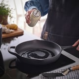 Person seasoning skillet with oil from bottle. Skillet on stove, knife nearby. Countertop has ingredients, plant.