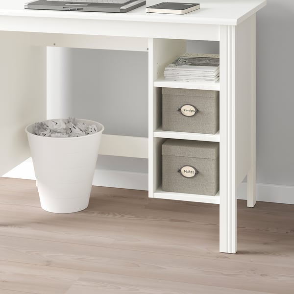 A white BRUSALI desk with adjustable shelves storing papers and boxes. A bin with crumbled paper sits beside it on a light wooden floor.