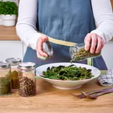 A person seasoning a salad with herbs from a CITRONHAJ spice jar, others with spices display, on a kitchen table.