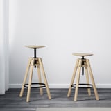 Two light wood adjustable bar stools against a white wall and a white curtain on a dark wood floor. Stools are shown with height adjustments.