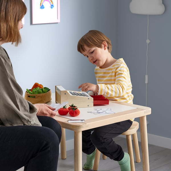 Child playing with DUKTIG toy cash register at table with play food. Boy wearing striped shirt, with adult nearby.