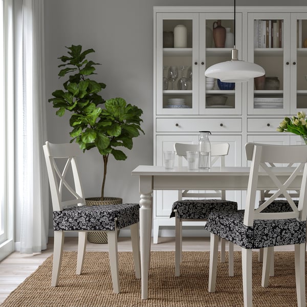 Modern dining table with floral chair pads, plant, and dish-filled cabinet.