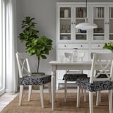 Modern dining table with floral chair pads, plant, and dish-filled cabinet.
