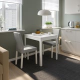 Modern white extendable dining set with two grey chairs, pendant lamp, dark rug, and city view window.