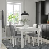 Modern extendable dining table with white top and grey chairs beside black cabinets, enhanced with bright lighting and plant decor.