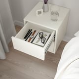 White nightstand with open drawer. Top: clock, water glass, purple flowers. Bed and curtains partly seen.
