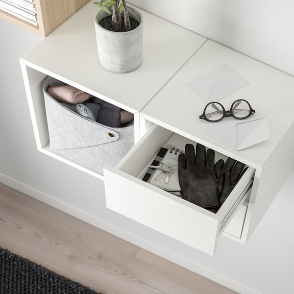 White wall-mounted storage unit, holding accessories. Drawer open, showing gloves, keys, and a notebook. Top has a plant and folded clothes.